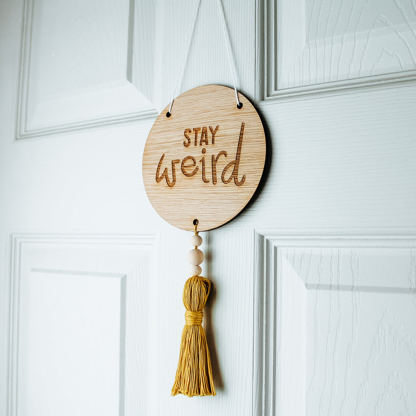 round wooden sign engraved with the phrase stay weird with a coloured tassel affixed to the bottom 