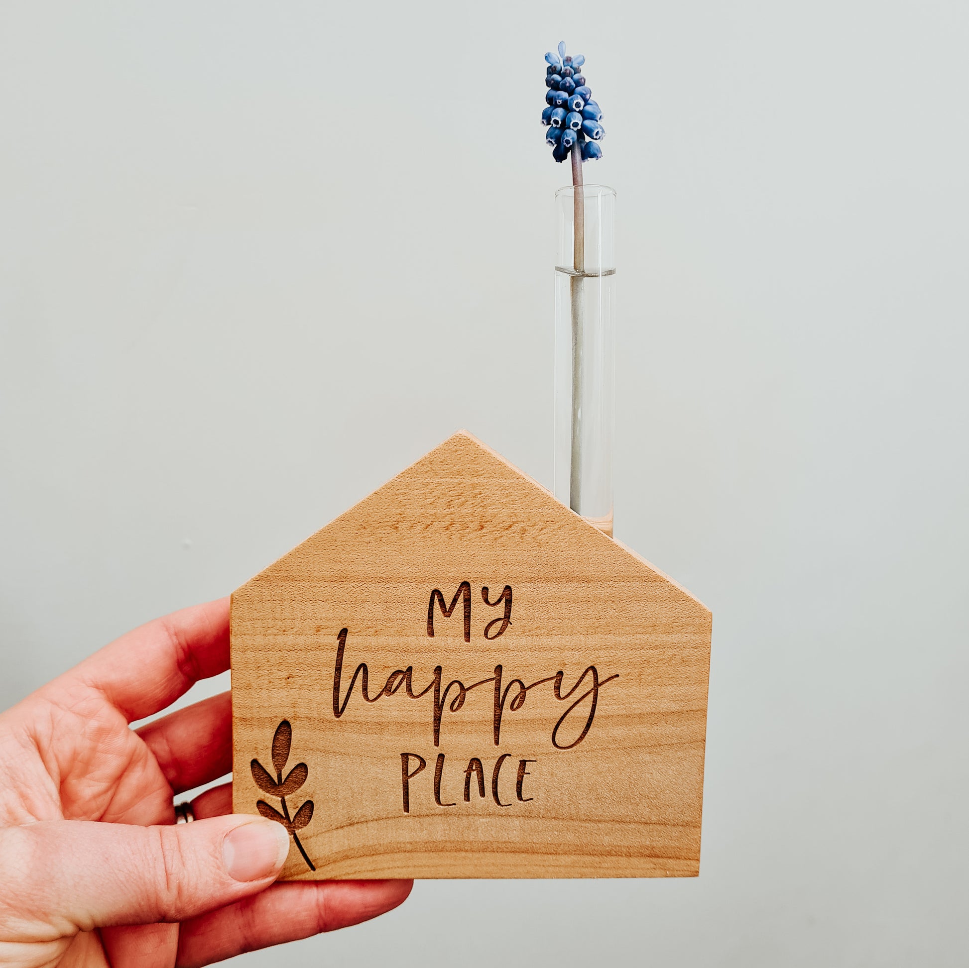 Wooden house shaped vase with test tube holding a single cut flower, engraved with the words my happy place